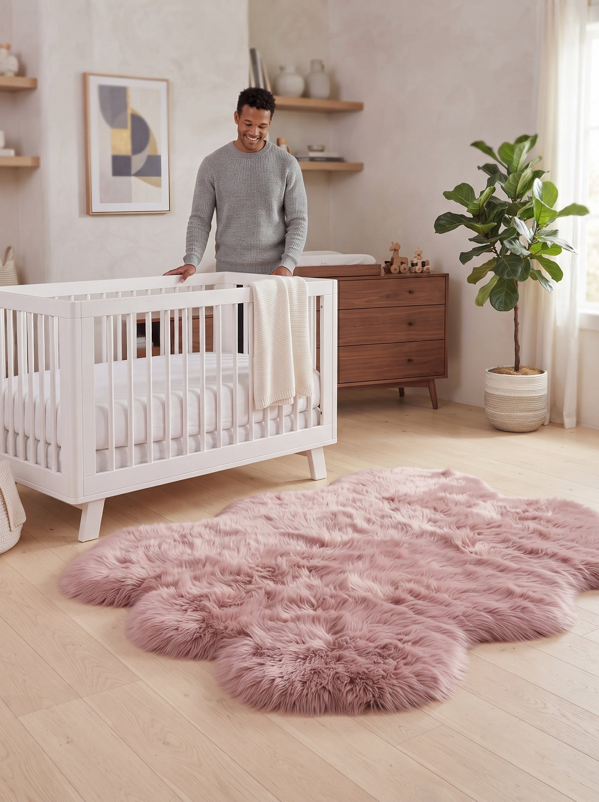A fluffy blush pink sheepskin-style area rug in an organic cloud shape on a light wood floor in a modern nursery. A man in a gray sweater stands behind a white wooden crib, and a wooden dresser, framed art, and a potted fiddle-leaf fig plant are visible in the room.