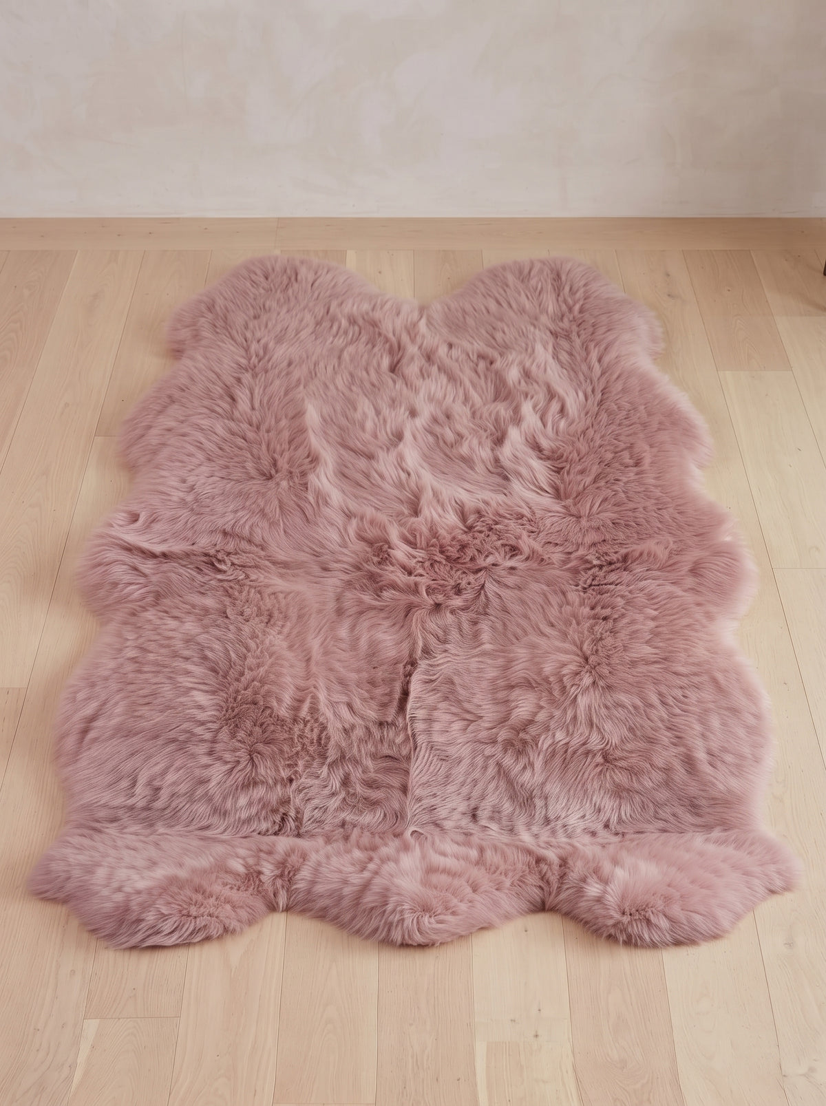 A dusty pink sheepskin rug with a natural curly texture, laid out on a light wood floor against a neutral beige wall.