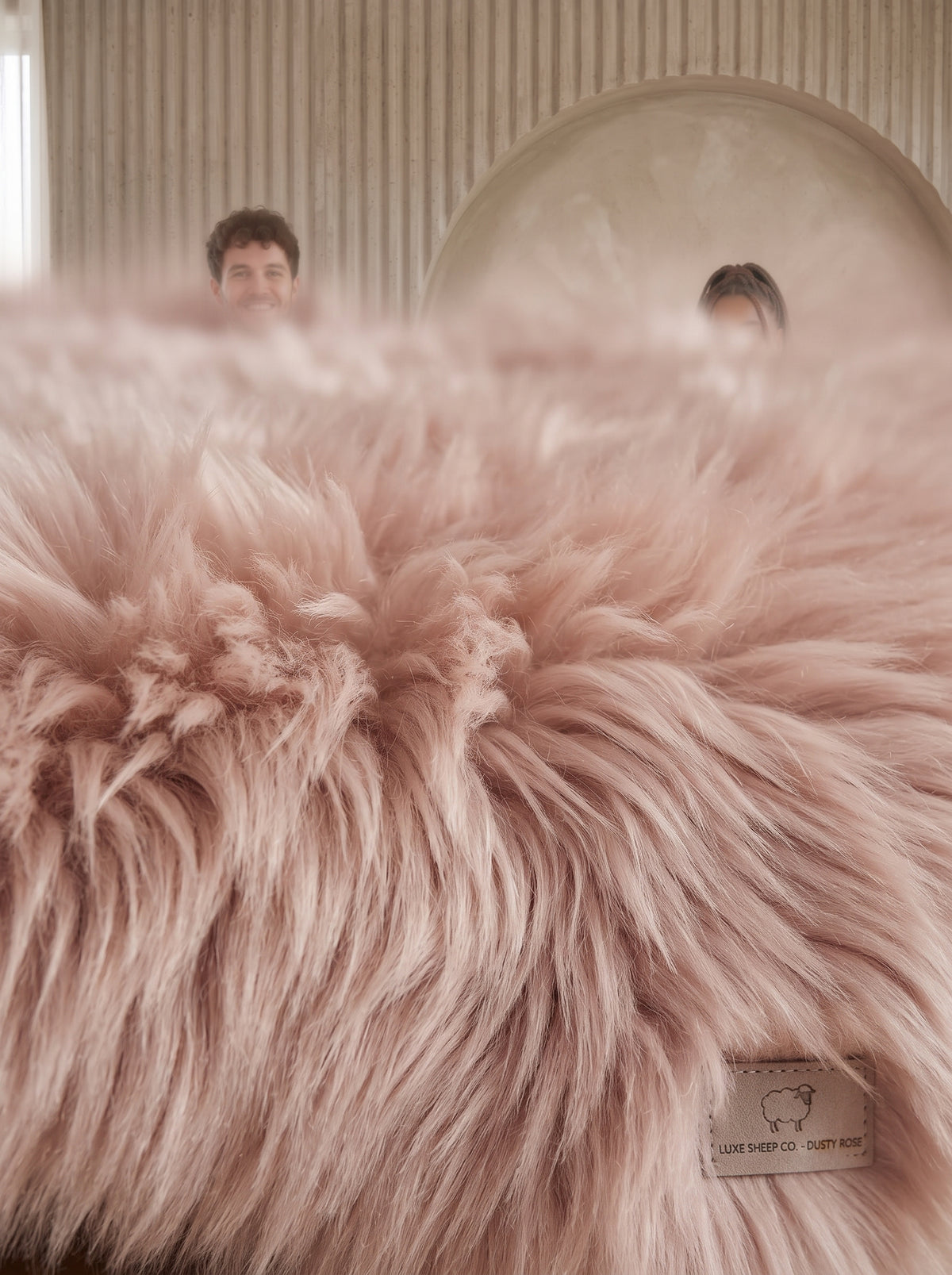 A close-up of a fluffy blush pink sheepskin or faux-sheepskin rug with a small sewn label in the lower right corner. In the softly blurred background, a person is visible behind a large oval headboard in a bedroom setting.