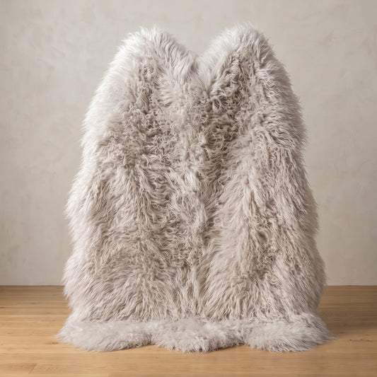 A fluffy faux-fur or sheepskin-style throw blanket in a light warm gray/beige tone, draped upright on a wooden floor against a textured beige wall.
