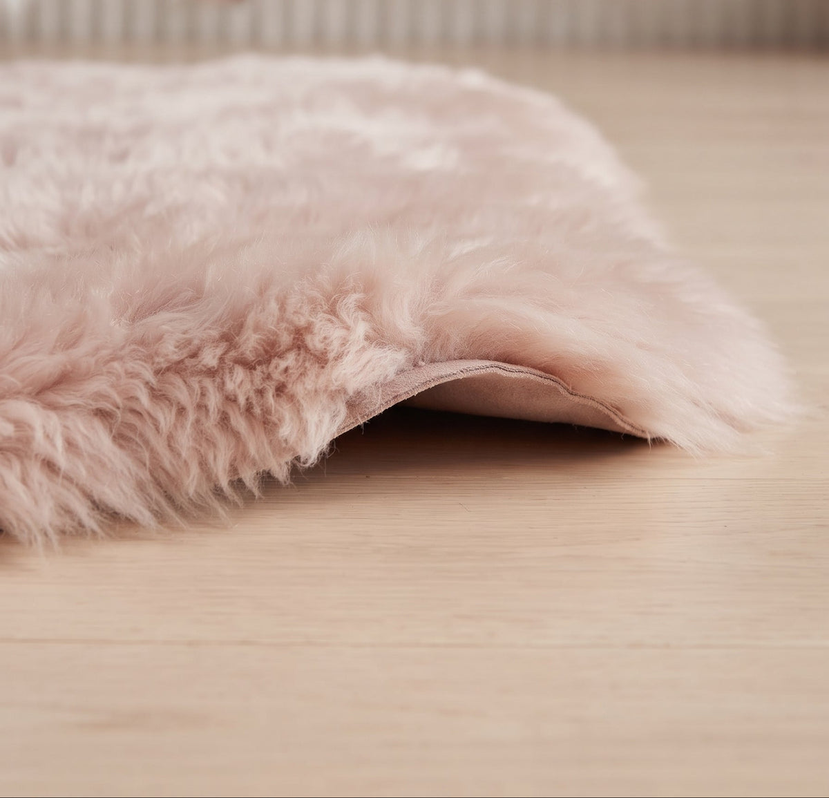 A pale blush pink fluffy sheepskin rug draped over a light wood floor, with a softly blurred man's face visible in the background near a cream-colored arched wall.