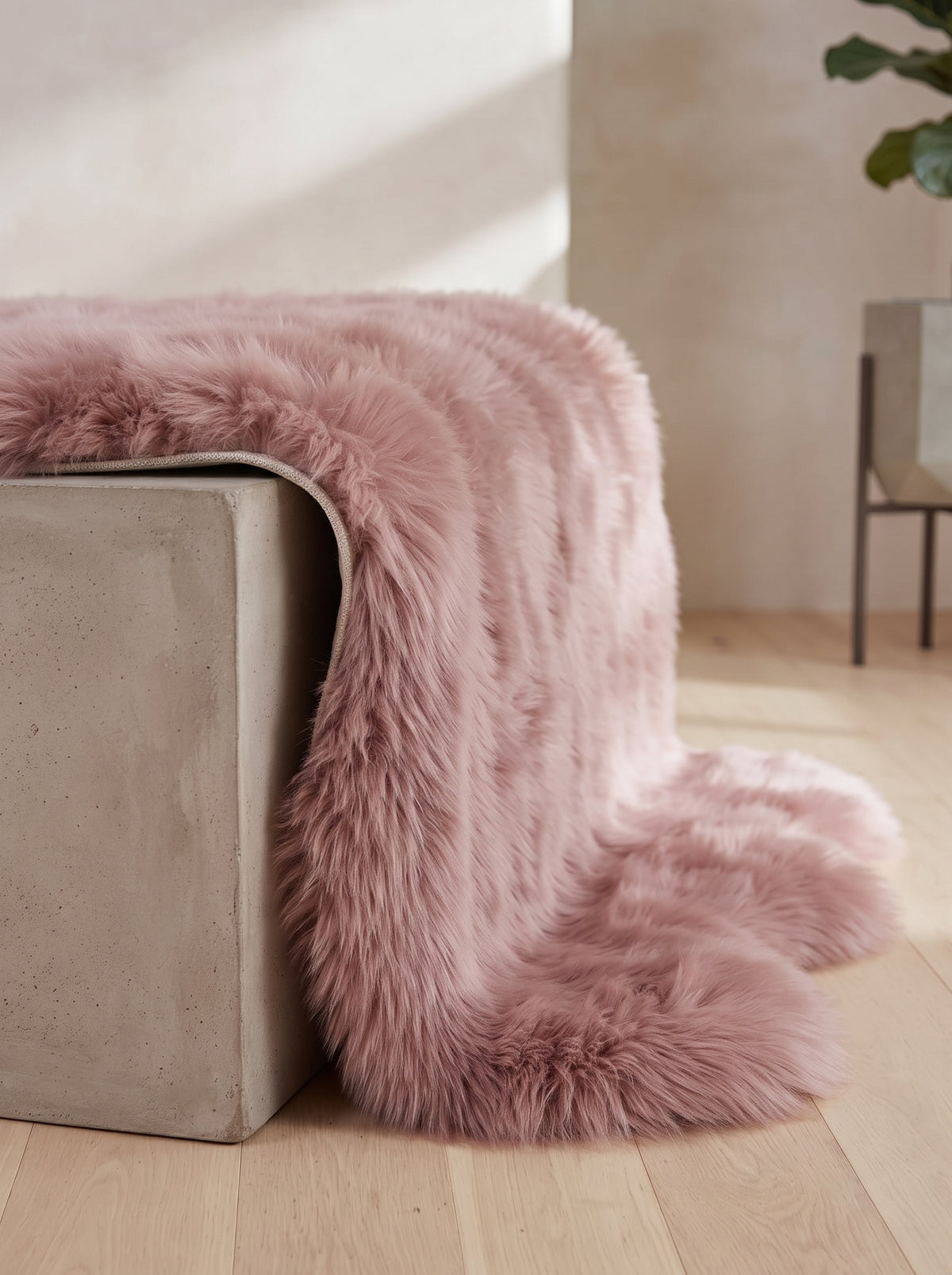 A dusty pink faux fur throw blanket draped over a concrete bench in a modern, sunlit room with light wood floors.